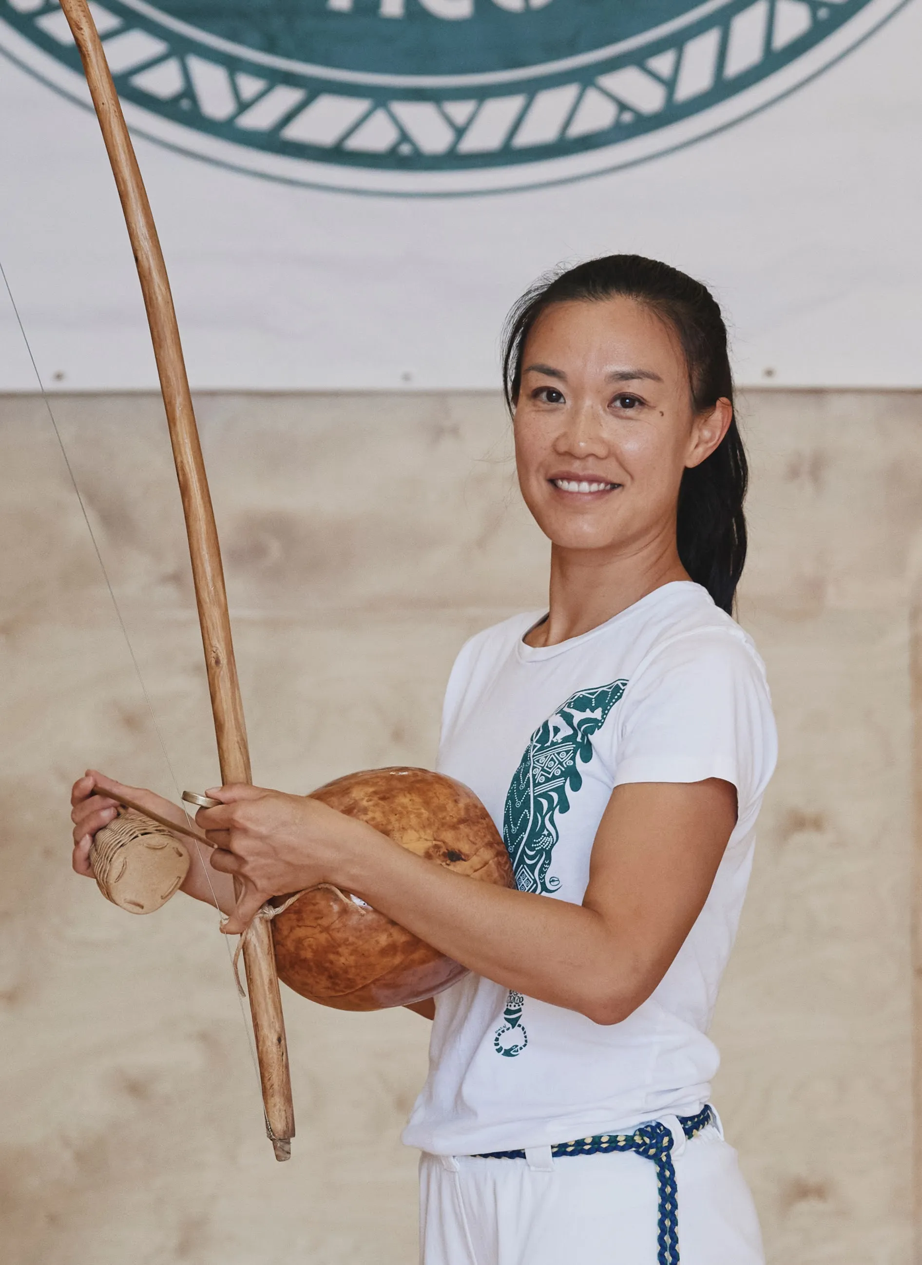 Professora Laurinha Rapadura holds a berimbau in her hands, in the studio with the Ilê Axé Capoeira logo partially visible