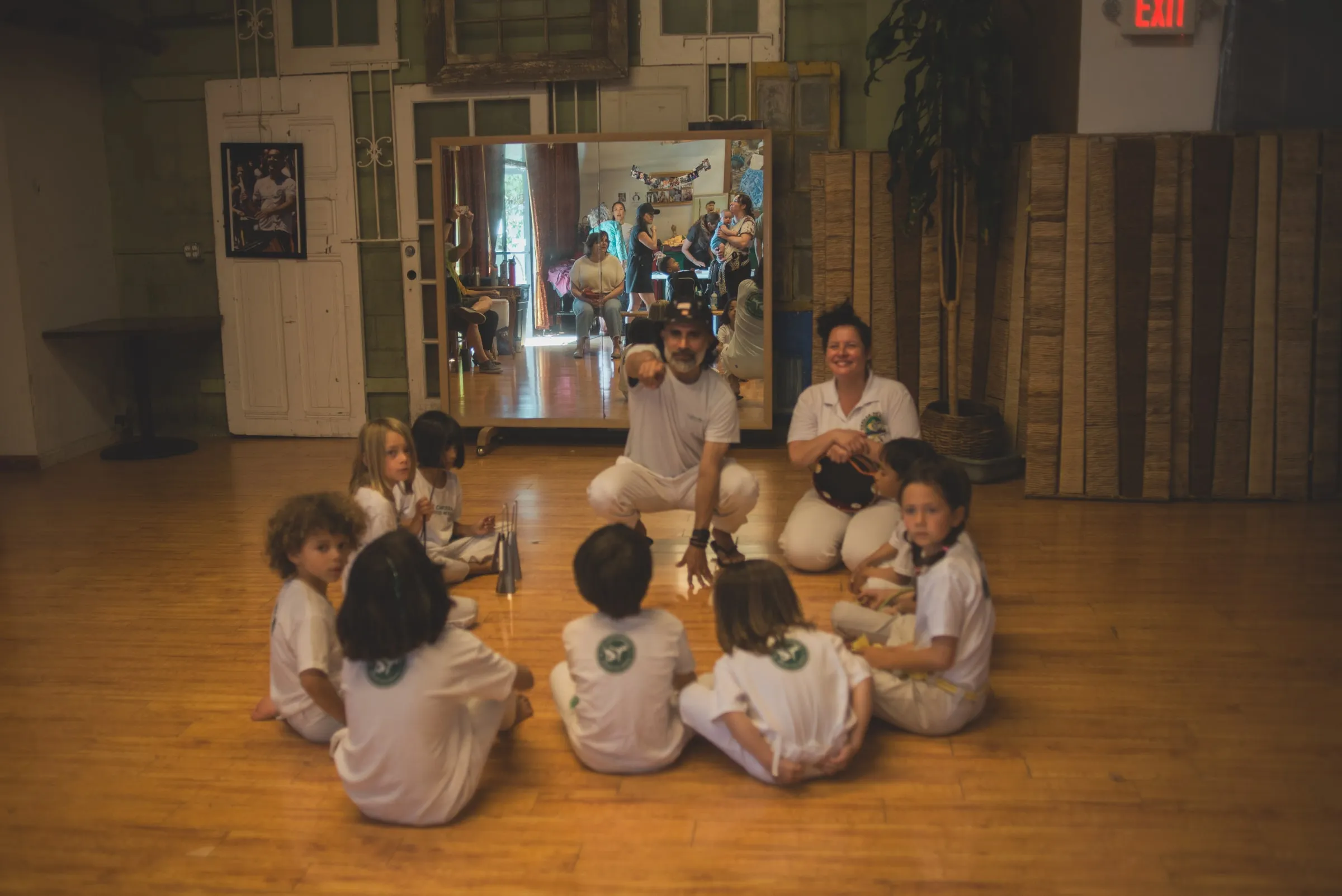 Children sit in a circle around two instructors during a kids capoeira class in a wood-floored studio.
