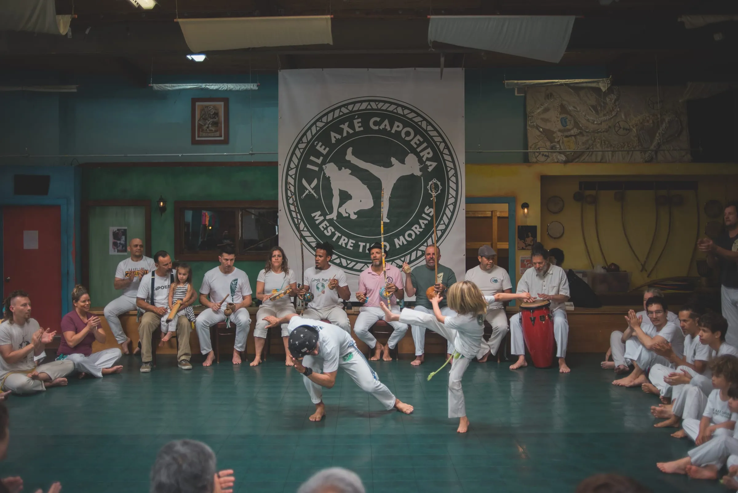 A roda fills a colorful hall as two capoeiristas play at the center and seated musicians perform beneath the round Ilê Axé Capoeira banner.