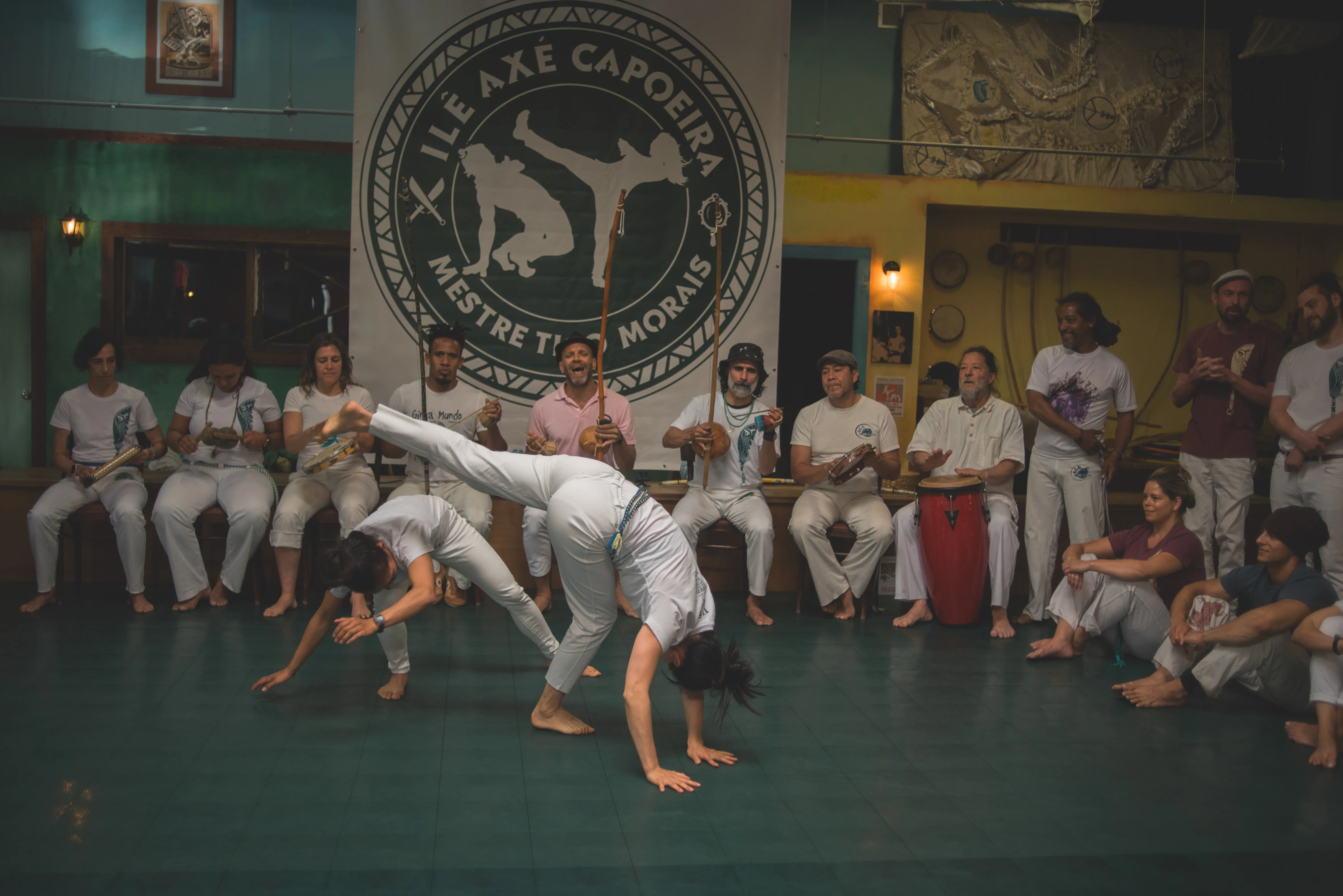 Two capoeiristas move through cartwheels in front of a seated bateria and gathered roda at an Ilê Axé event.