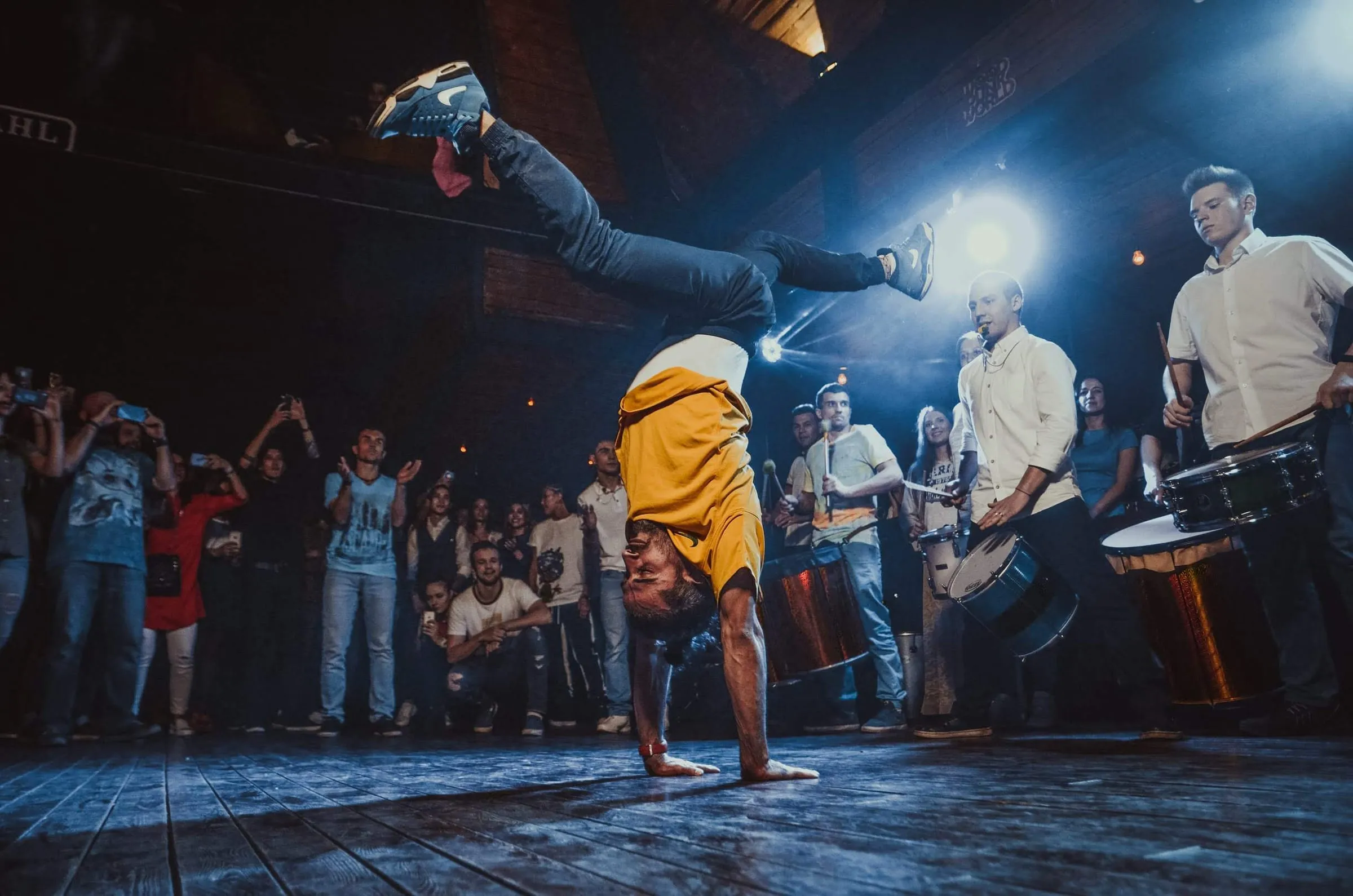 Mestre Tico holds a one-armed handstand on a dark stage while musicians perform and a crowd watches under stage lights.