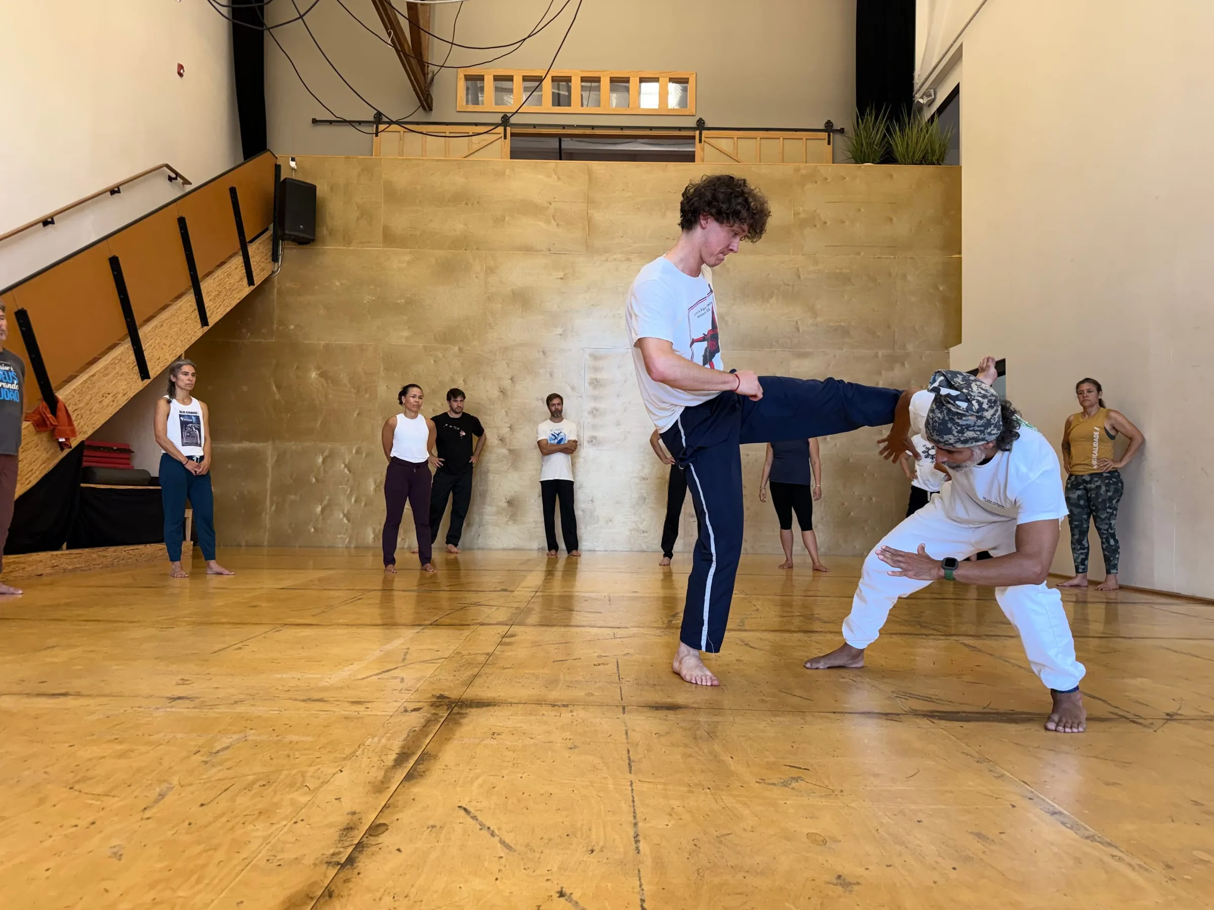 An instructor coaches a student mid-kick during an adult class while other students watch from the edge of a bright wood-panelled studio.