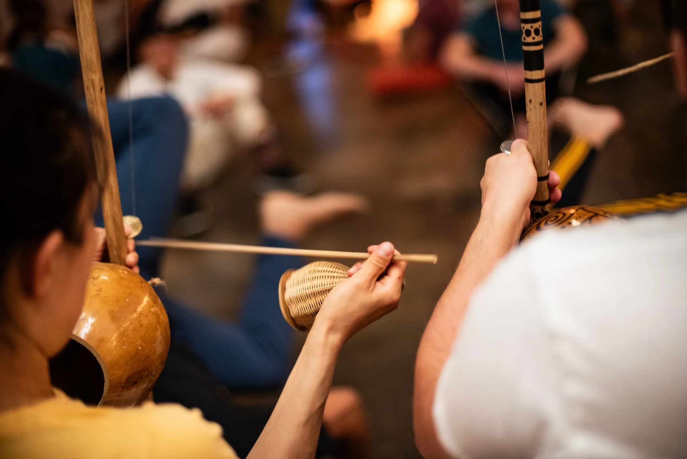 Close view of hands playing a berimbau: one hand holds the caxixi rattle and striking stick while the gourd and wire of the instrument blur into a warm room.