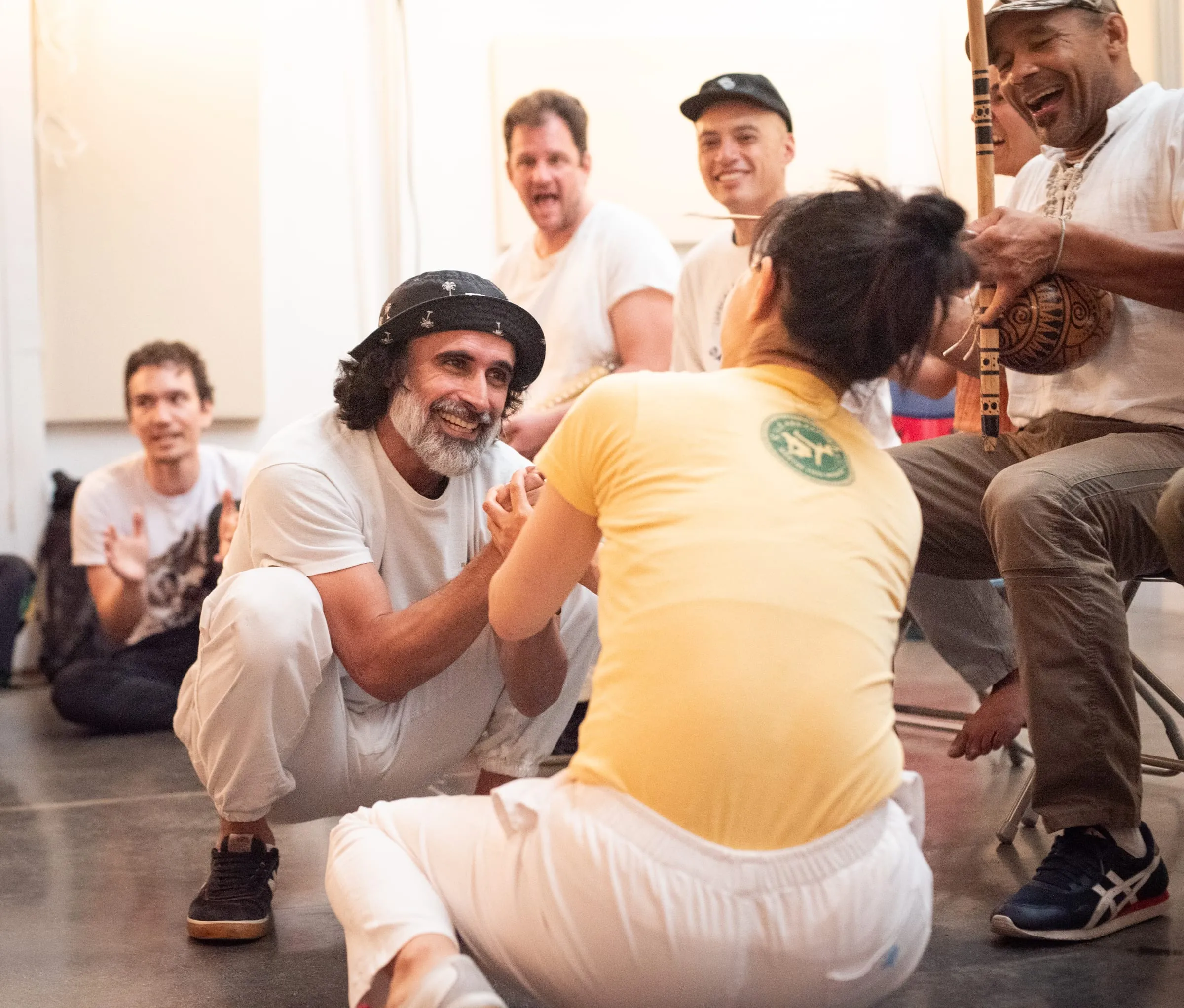 Two capoeiristas clasp hands and smile at the center of the roda while musicians and students look on.