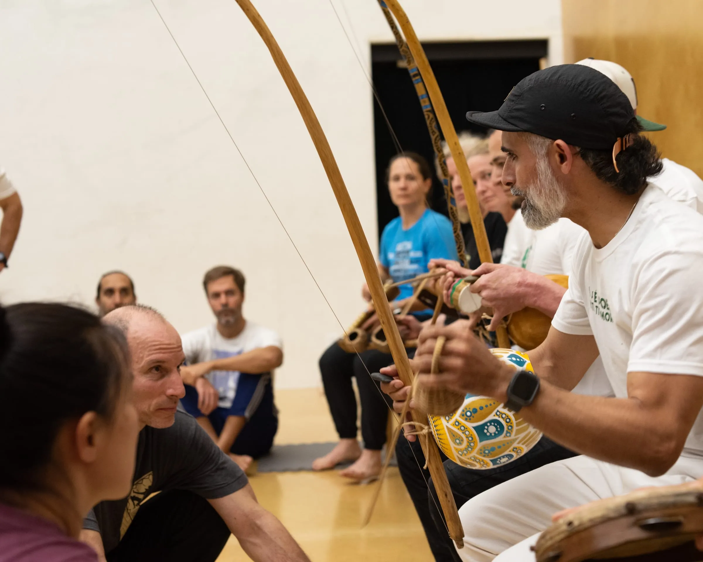 An instructor holds a berimbau upright and explains its rhythm while students seated around the room listen closely.