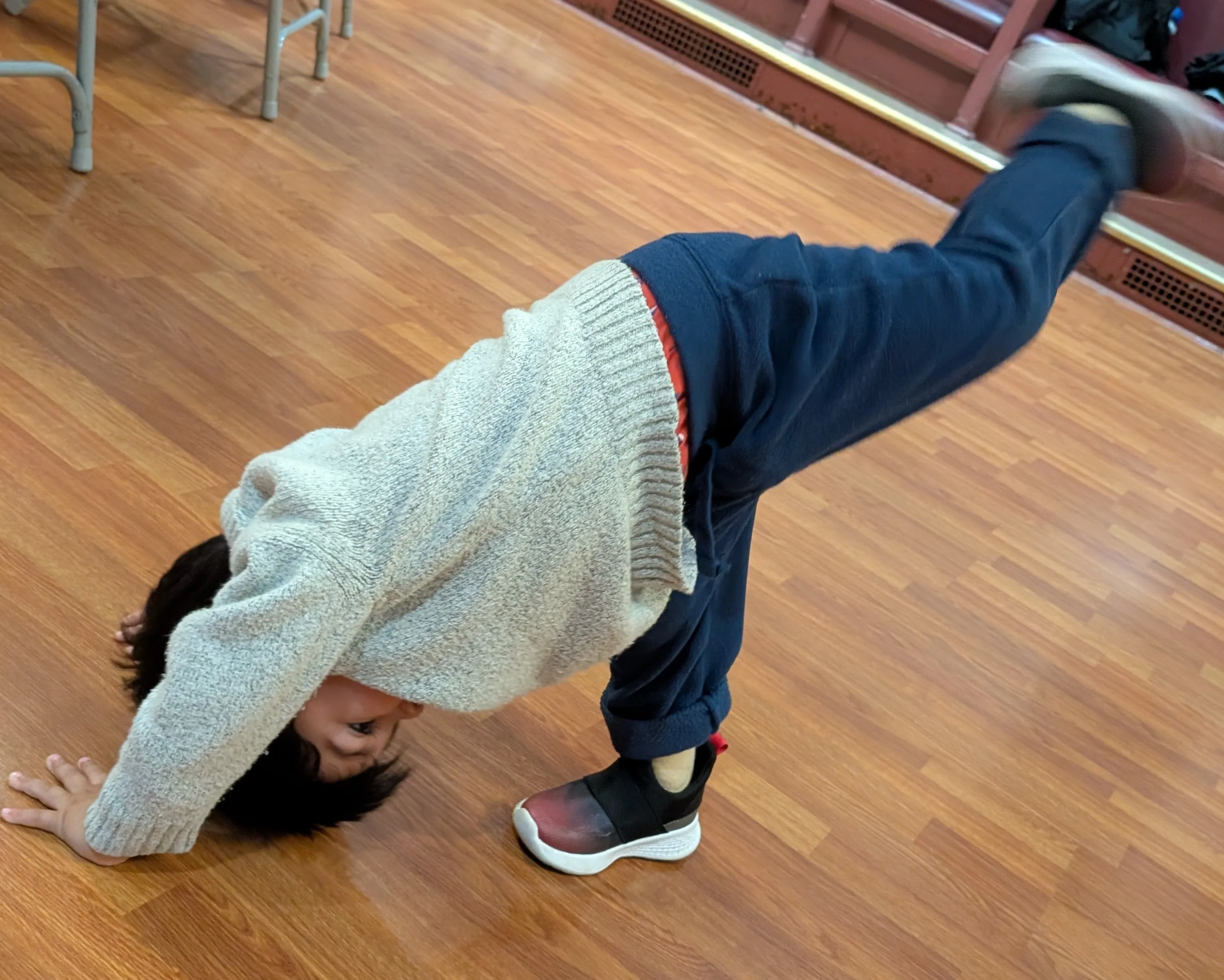 A young child practices a playful hands-on-the-floor movement with one leg raised in a wood-floored room.